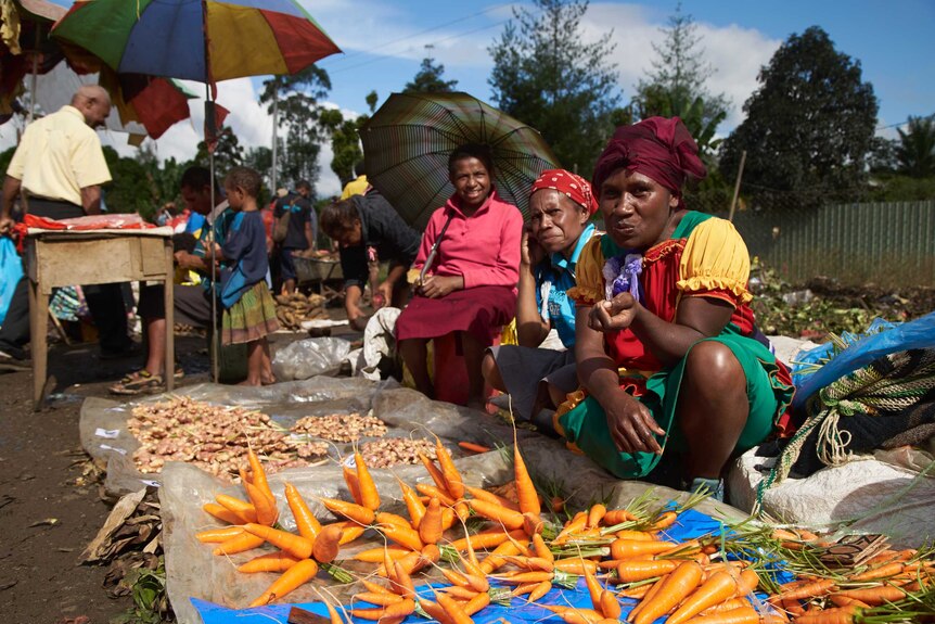 Three PNG women sit on the ground next to sheets of plastic laid out, selling carrots and ginger. One holds an umbrella.