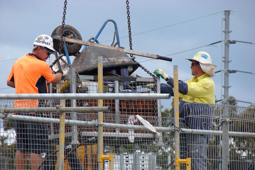 Two constructions unload items from a steel cage being lowered by a crane, including an upside down wheelbarrow