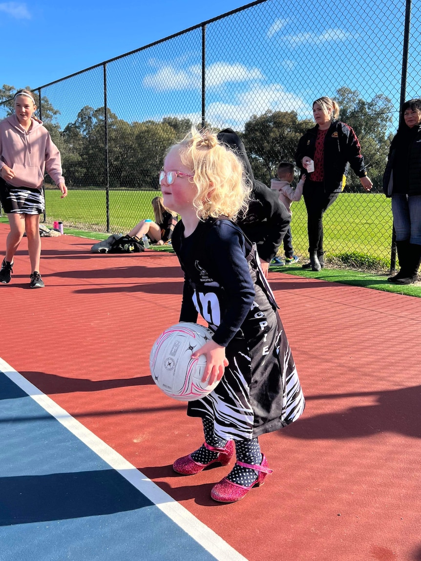 young girl with blonde curly hair and black netball uniform, holding a white ball, knees bent, pink heel shoes