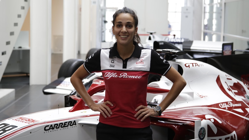 Krystina Emmanouilides stands in front of formula one cars with her hands on her hips.