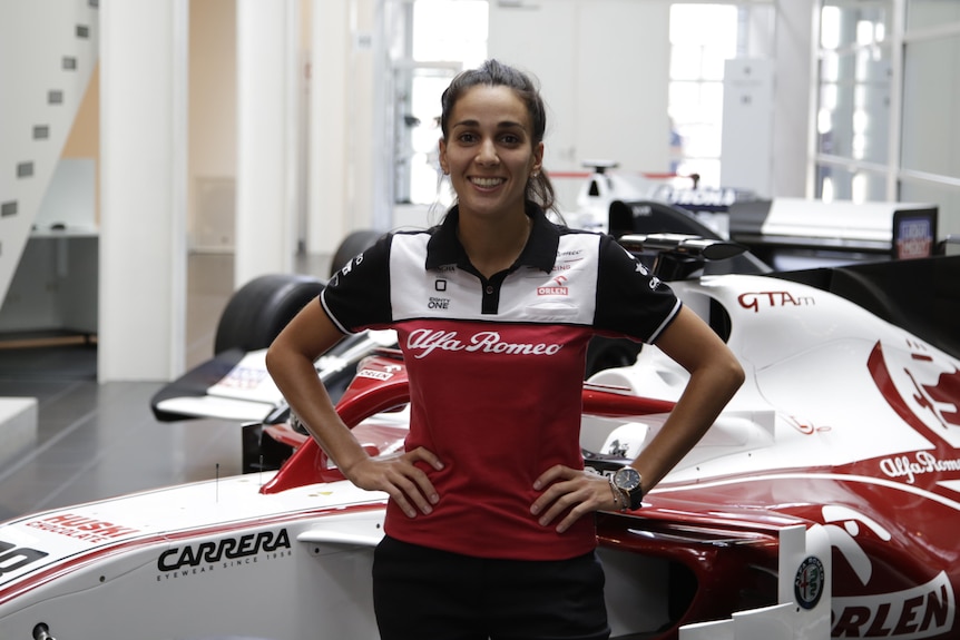 Krystina Emmanouilides stands in front of formula one cars with her hands on her hips.