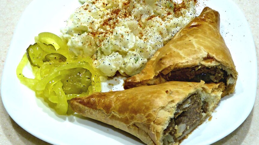 A Cornish pasty on a plate with some pickles and potato salad.