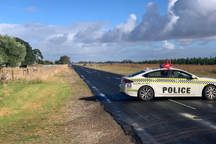 A police car across a country road to block it, fields and figures around.