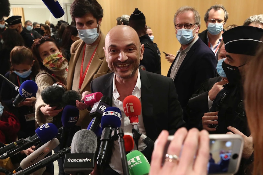 A man in a suit smiles as he talks to a large pack of media that has surrounded him while holding mics and cameras.