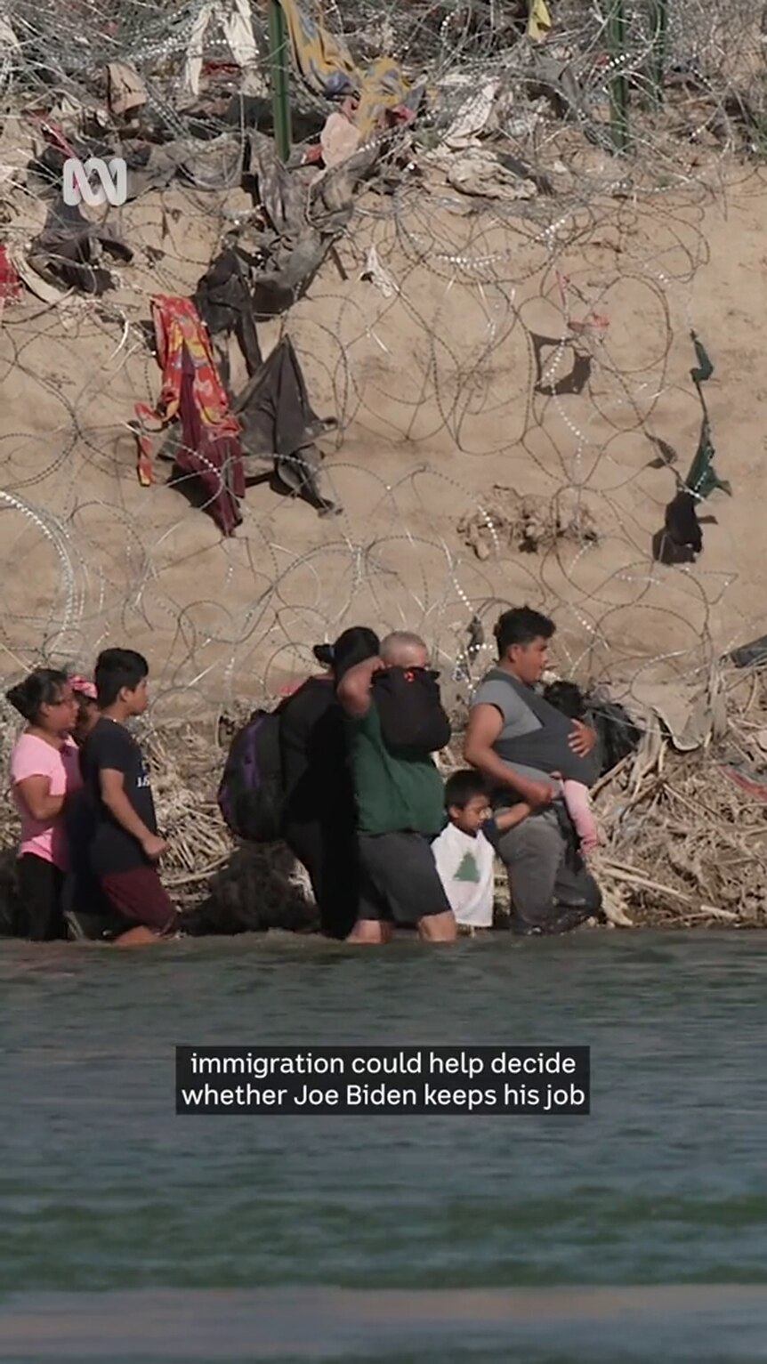 Brown-skinned family wades through water on the edge of a river bank covered in razor wire