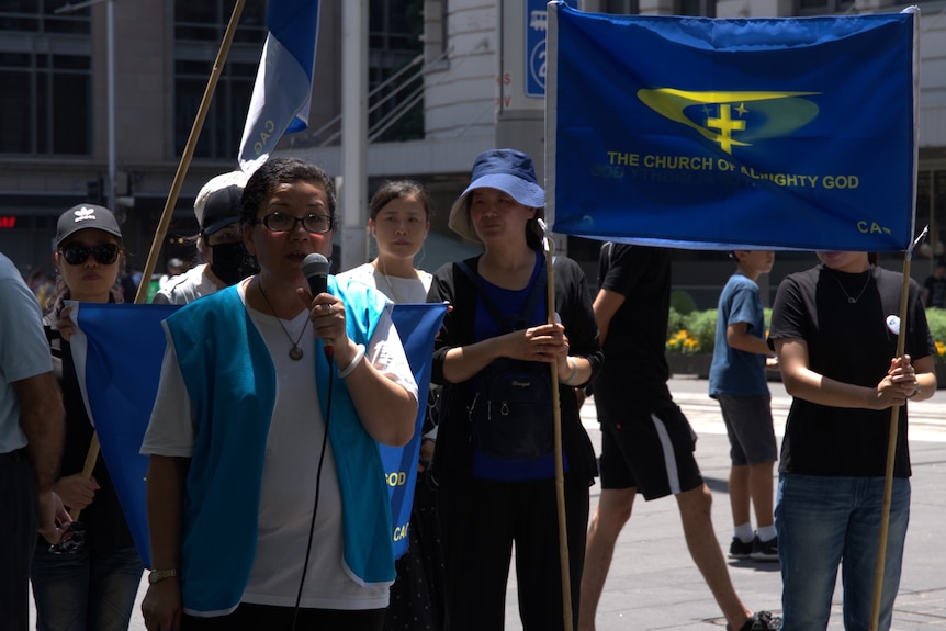 A woman in blue holding a microphone in front of a crowd with a flag.