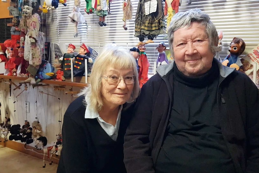 An older couple pose in front of a large collection of puppets