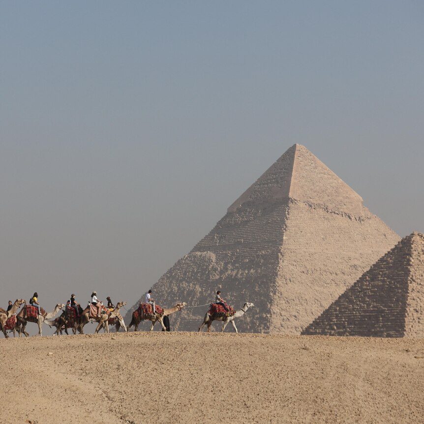 People ride horses between one large and one smaller pyramid.