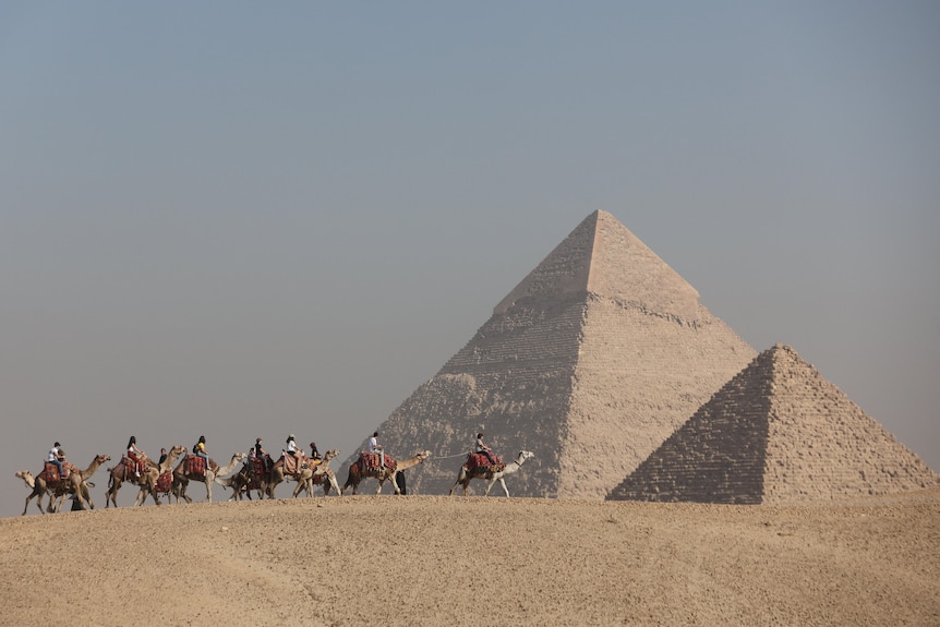 People ride horses between one large and one smaller pyramid.