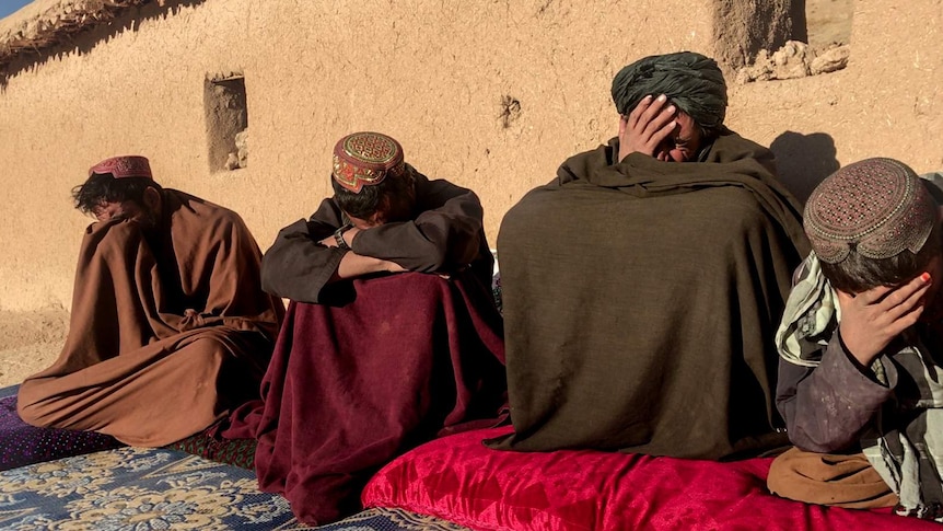 Three men and a young boy, sitting in robes and visibly grief stricken, sitting in front of a compound.