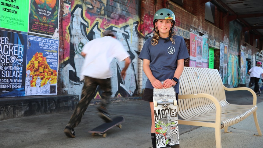 A young girl named Taylah Payne smiles while holding a skateboard as another person skates behind her.