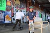 A young girl named Taylah Payne smiles while holding a skateboard as another person skates behind her.