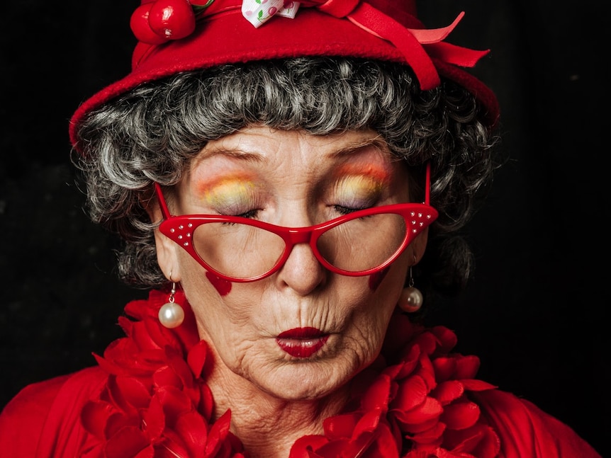 Older woman with grey hair, vibrant red hat, top and glasses, rainbow coloured eyeshadow. Eyes closed and 'kiss' mouth.
