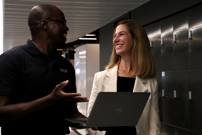 A woman with should length dark blond hair wearing a white suit jacket talks to a man with a black polo shirt holding a laptop