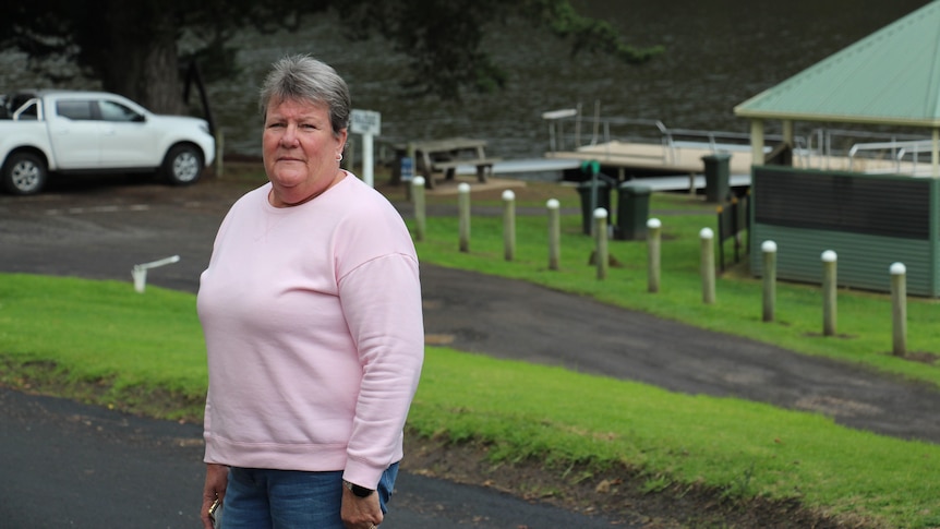 A short-haired woman in a pink jumper stands, a pontoon, pavilion and river behind her