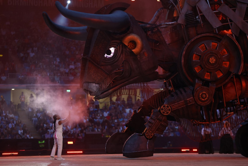 a dancer holds her hand up towards the face of a giant 10m tall mechanical bull in the middle of a stadium