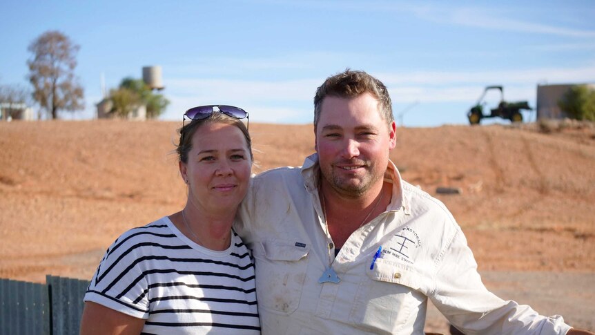 A woman and a man stand in front of a corrugated iron fence with a red dirt-covered hill in the background.