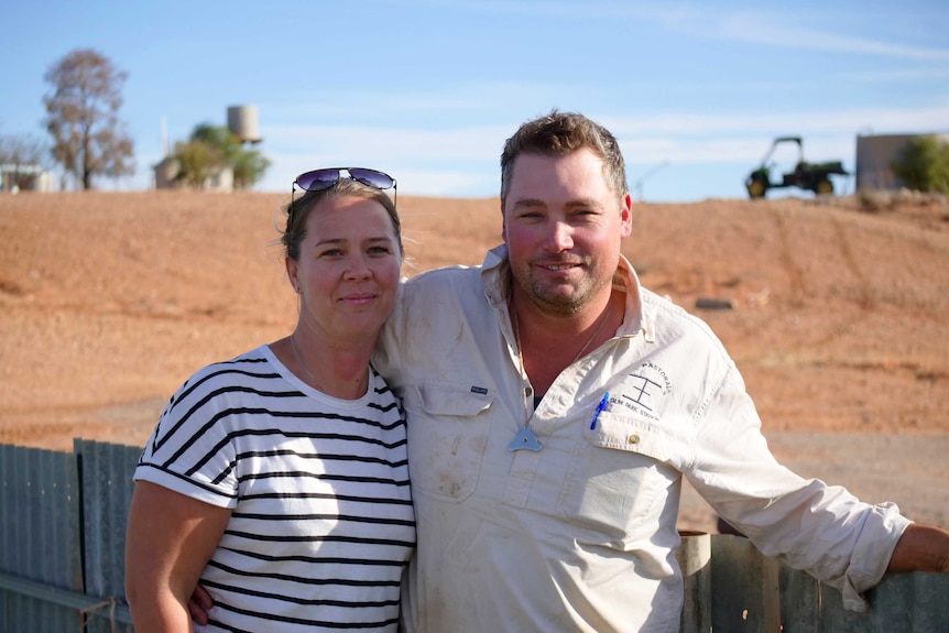 A woman and a man stand in front of a corrugated iron fence with a red dirt-covered hill in the background.