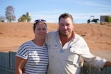 A woman and a man stand in front of a corrugated iron fence with a red dirt-covered hill in the background.