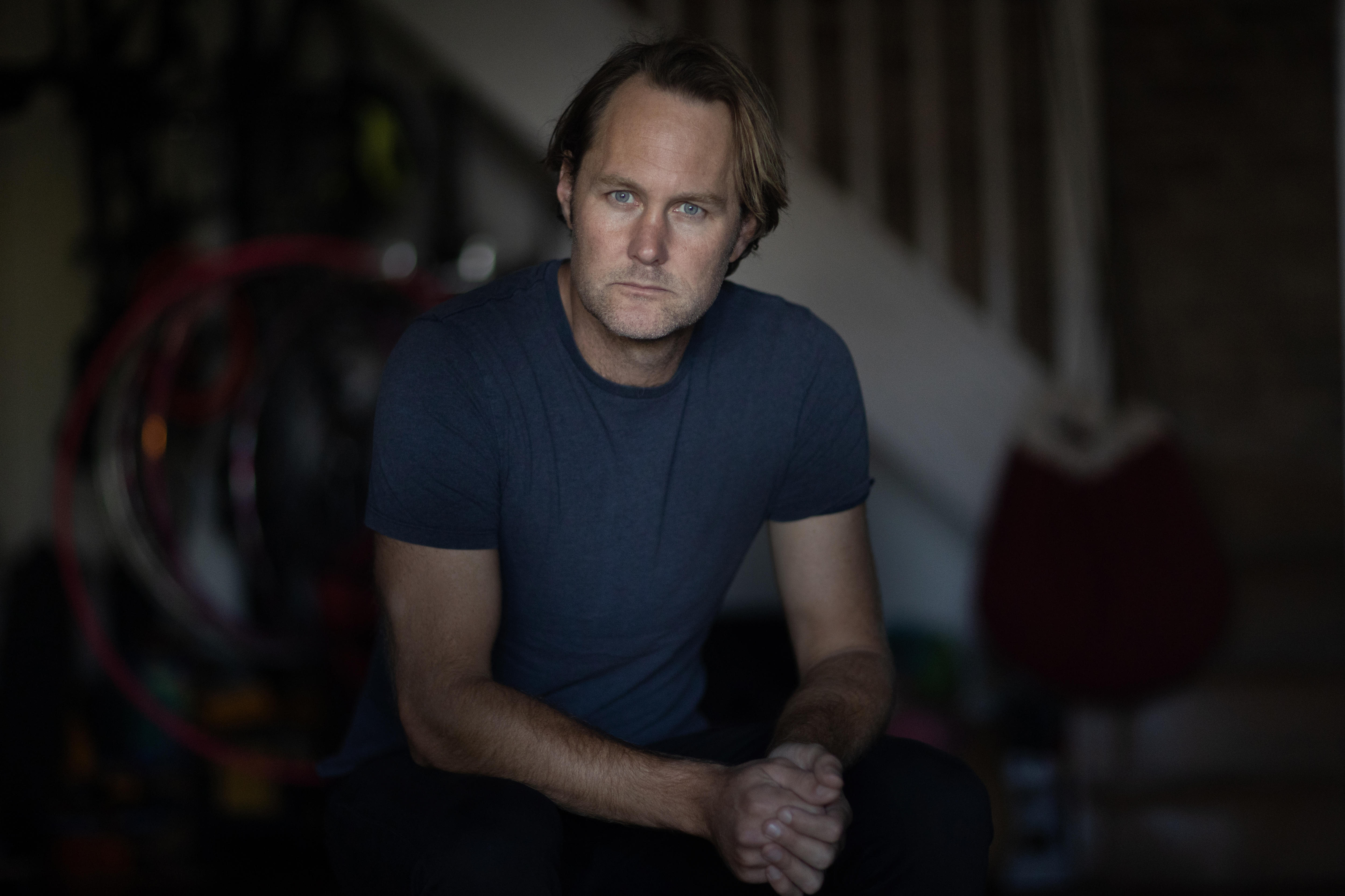 Una imagen oscura de un hombre vestido con una camiseta azul y mirando al frente con las manos cruzadas.