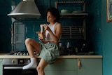 A brown woman wearing boxer shorts, a singlet and socks sits on a kitchen benchtop and eats noodles from a box.