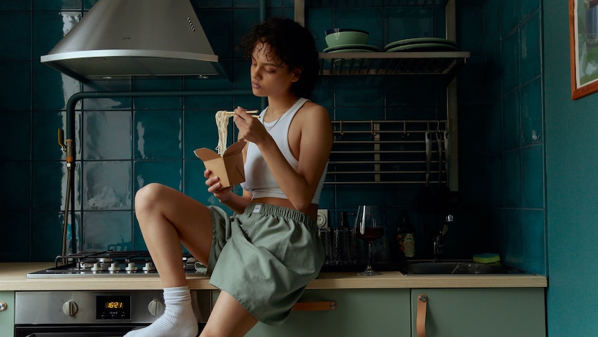 A brown woman wearing boxer shorts, a singlet and socks sits on a kitchen benchtop and eats noodles from a box.
