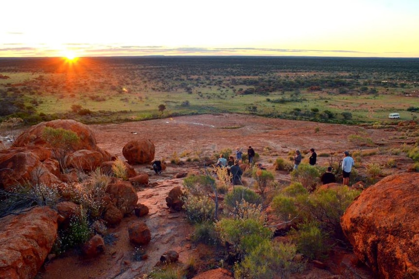People stand and sit at red rock earth overlooking a grassy plain as the sunsets in background
