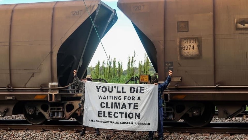 A protest sign reads "You'll die waiting for a climate election" between two coal railway wagons.