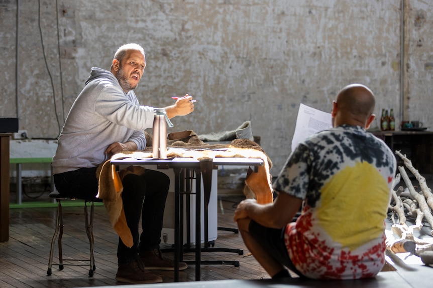A man in a grey hoodie sits at a table holding a pen, giving instructions to a man wearing an Aboriginal Australian shirt.