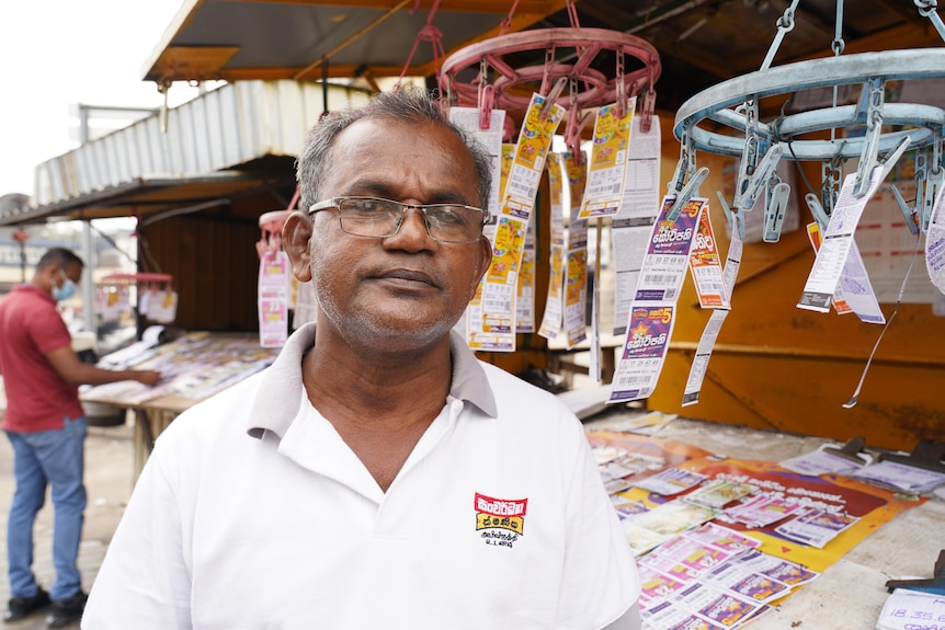 A middle-aged man in a polo shirt stands next to a street stall with lotto tickets displayed on laundry hangers