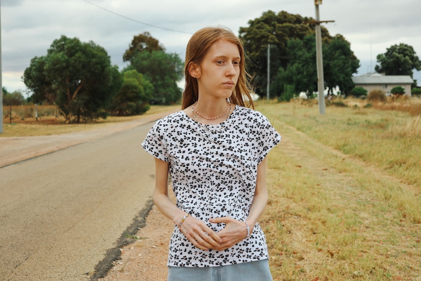 A young woman walks alongside a country road. Her expression is troubled and her arms are very thin.