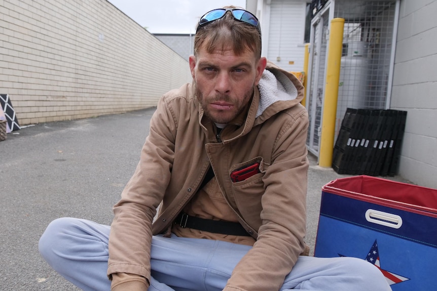 A man in a tan hoodie and jeans sits on the ground in a laneway area.