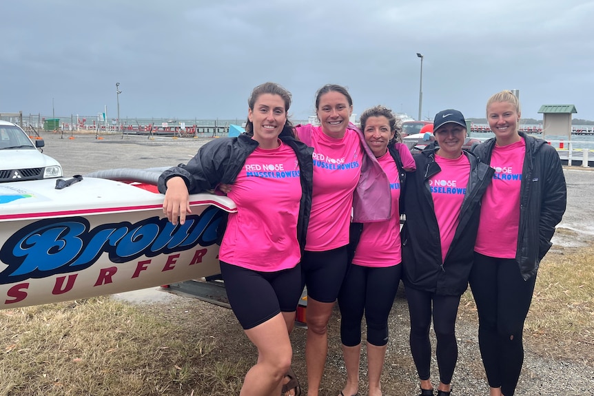 Five women wearing matching pink T-shirts stand in front of a boat near the shore