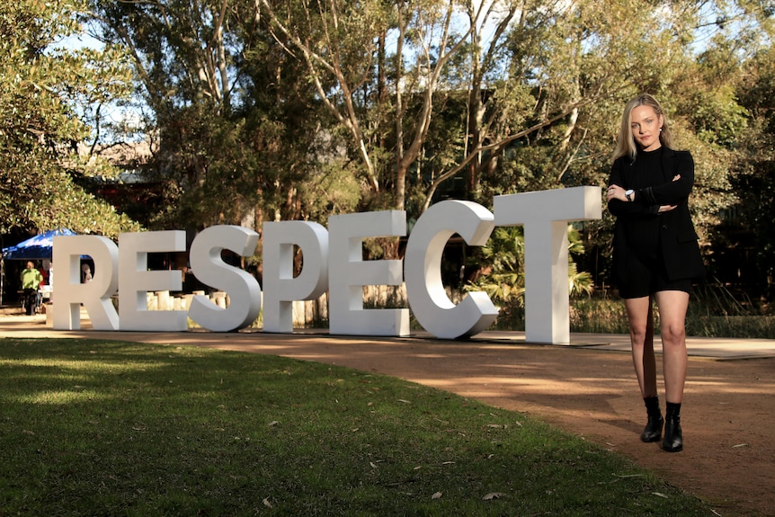 A young woman wearing dark clothes stands in front of a giant "respect" sign.
