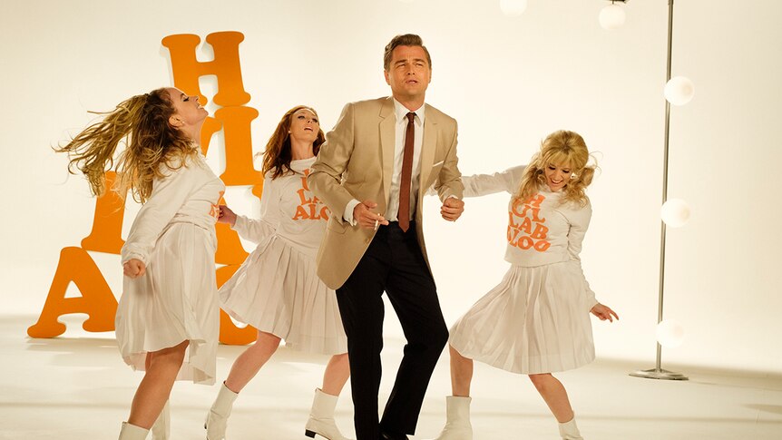 A man dressed in a suit holding cigarette dances on a film set with orange letters with three female dancers.