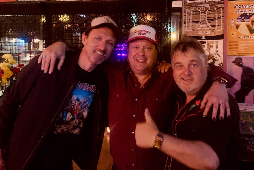 Three men standing inside a music venue surrounded by band posters