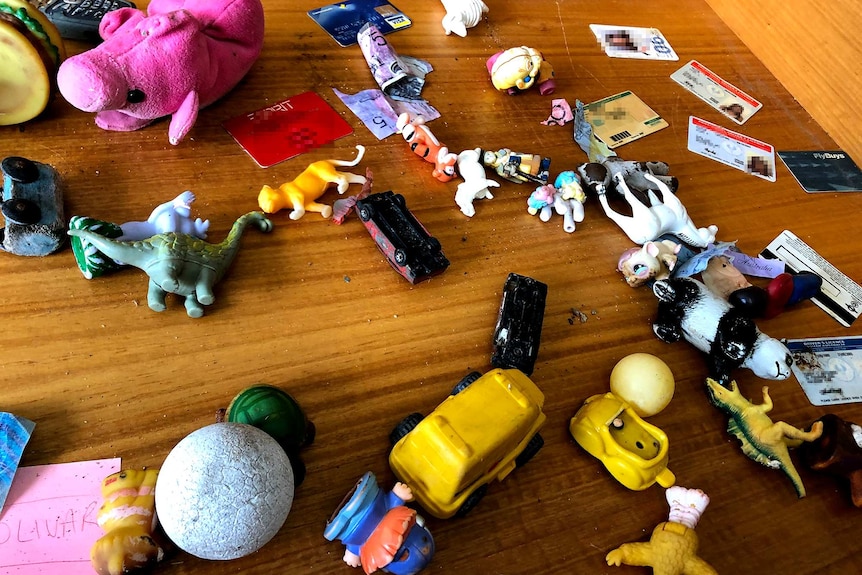 Stuffed toys, toy cars, money and drivers' licences sit upon a wooden bench.