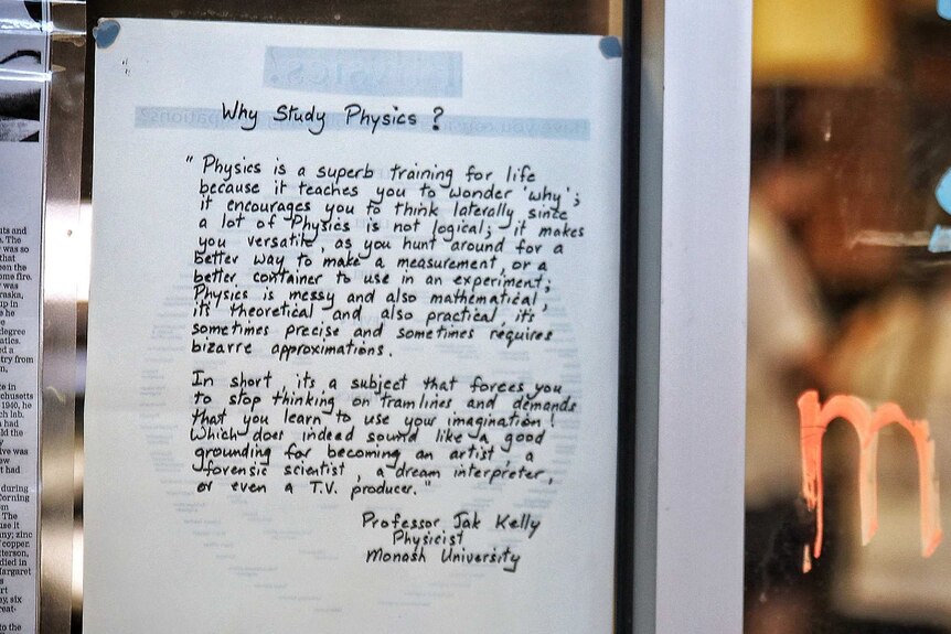 A written 'Why study physics' quote on a piece of white paper stuck on a glass wall in a classroom.