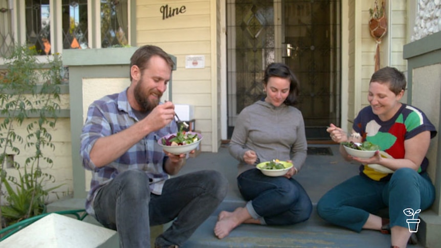 Man and two women sitting on front steps of house eating bowls of salad