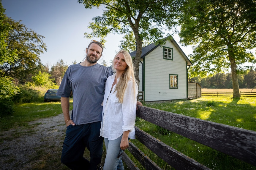 A couple stand next to a wooden fence in front of their house in Sweden.