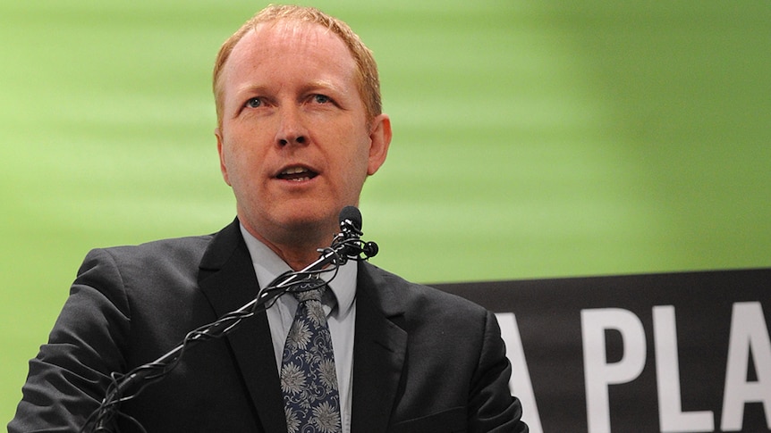 Man in suit giving a speech in front of a Greens banner.