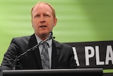 Man in suit giving a speech in front of a Greens banner.