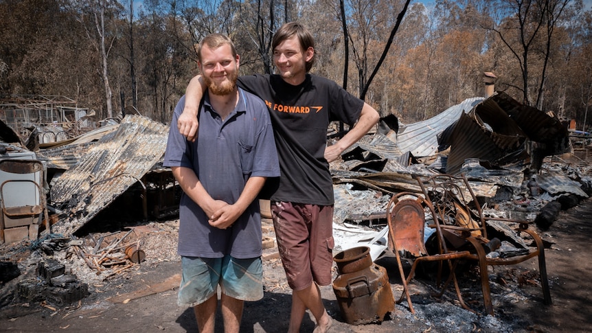 Two young men standing in front of a house burnt to the ground with sheets of twisted corrugated iron.