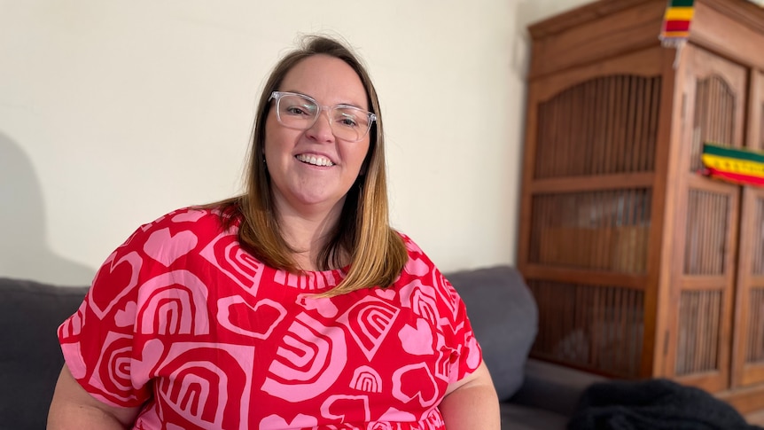 A woman wearing glasses and a pink and red dress. She is smiling and sitting on a couch.