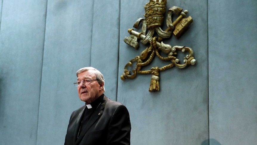 George Pell stands before a microphone, hands crossed over each other.