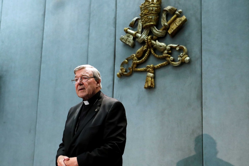 George Pell stands before a microphone, hands crossed over each other.