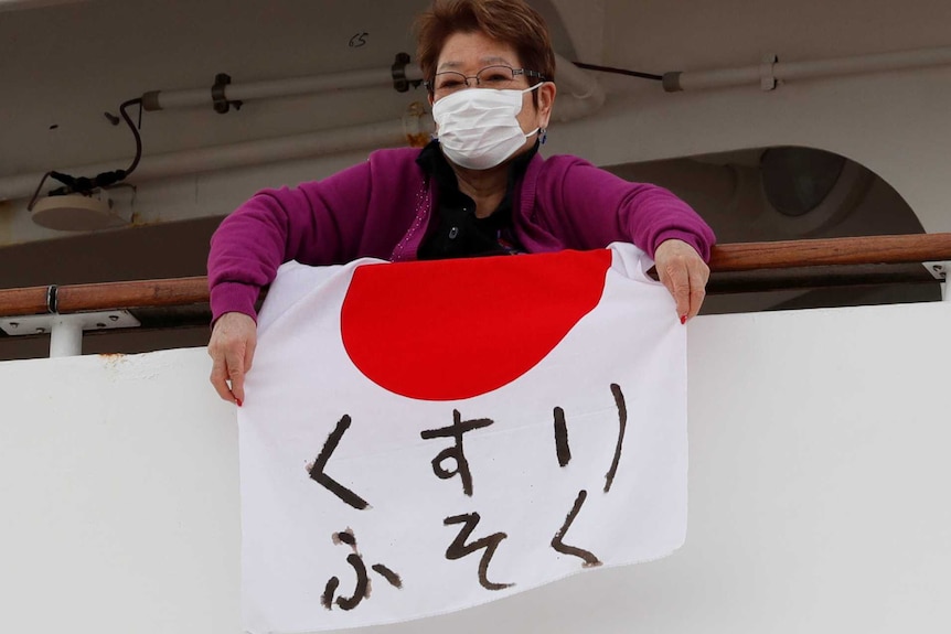 A woman wearing a mask hangs a Japanese flag with writing off the rail of a cruise ship.
