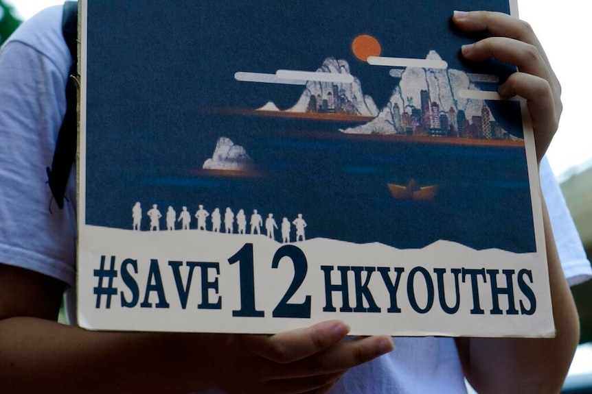 A person holds a placard that reads #Save12HKyouths with an illustration of a boat and Hong Kong's skyline.