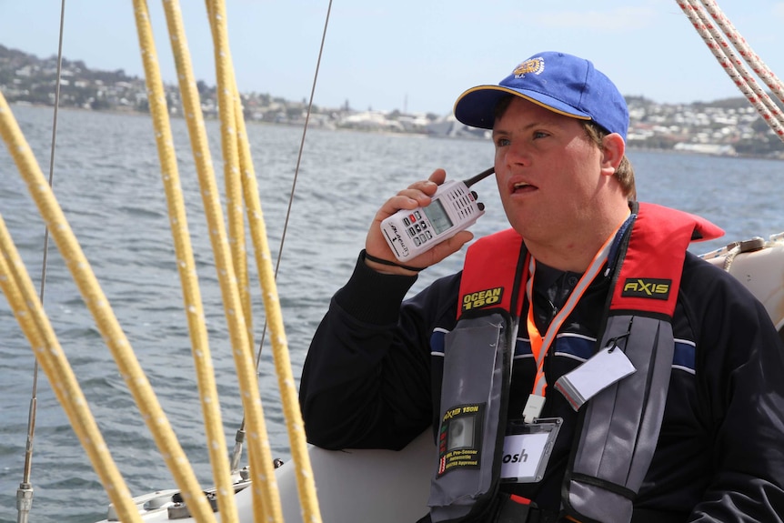 A man with Down syndrome holding a walkie talkie to his mouth, sitting on a sailing boat on the ocean.