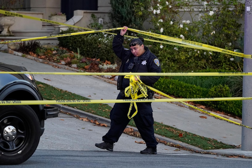 A police officer rolls out more yellow tape on the closed street below the home of Paul Pelosi.